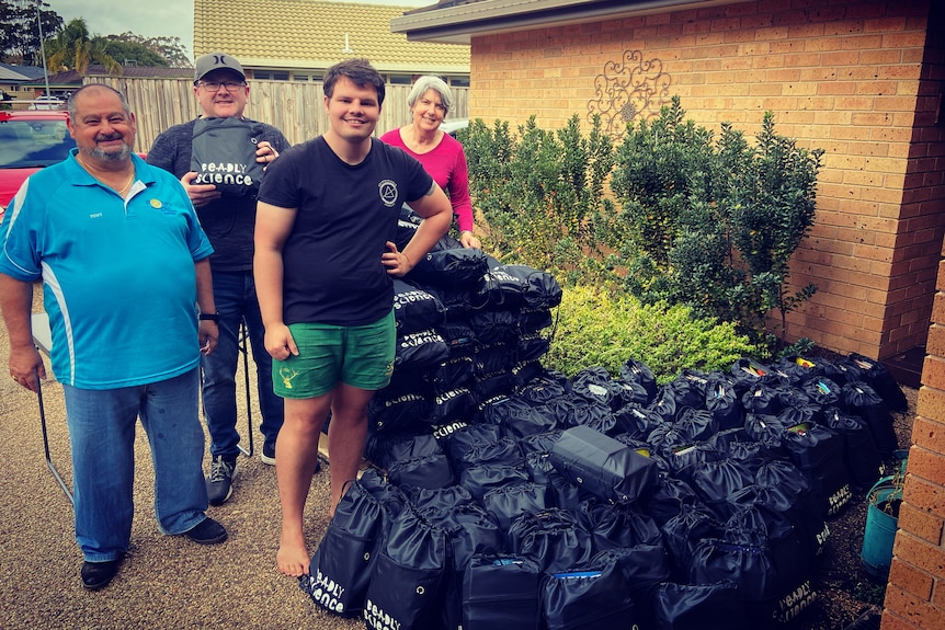 A smiling, barefoot man stands alongside two other men and a woman, with dozens of plastic bags beside them.