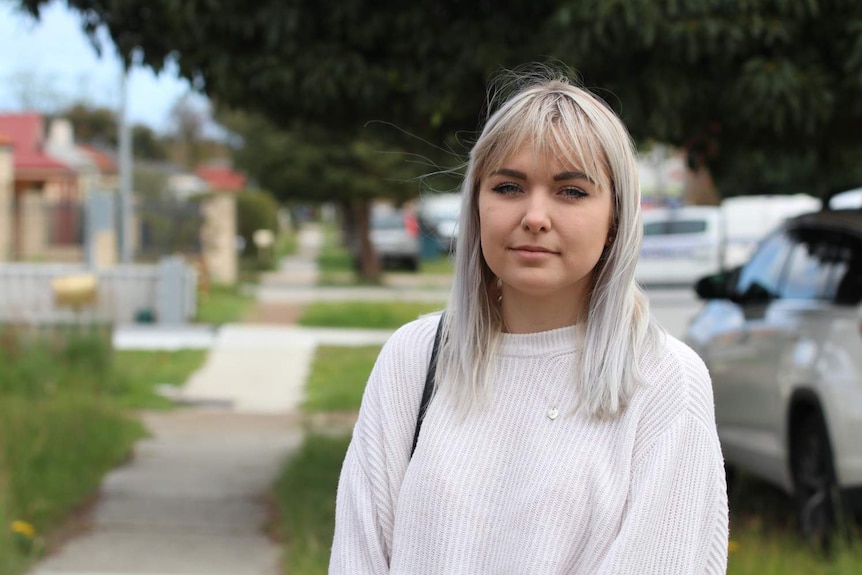 Hailie wearing a white jumper, standing near a footpath under a tree on a suburban street.