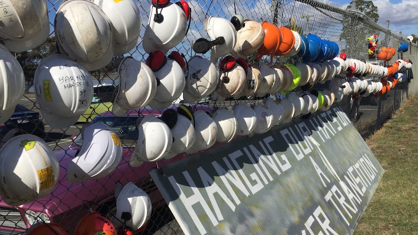 Dozens of hard hats hang on a cyclone fence, above a sign reading "hanging out hats on a worker transition".