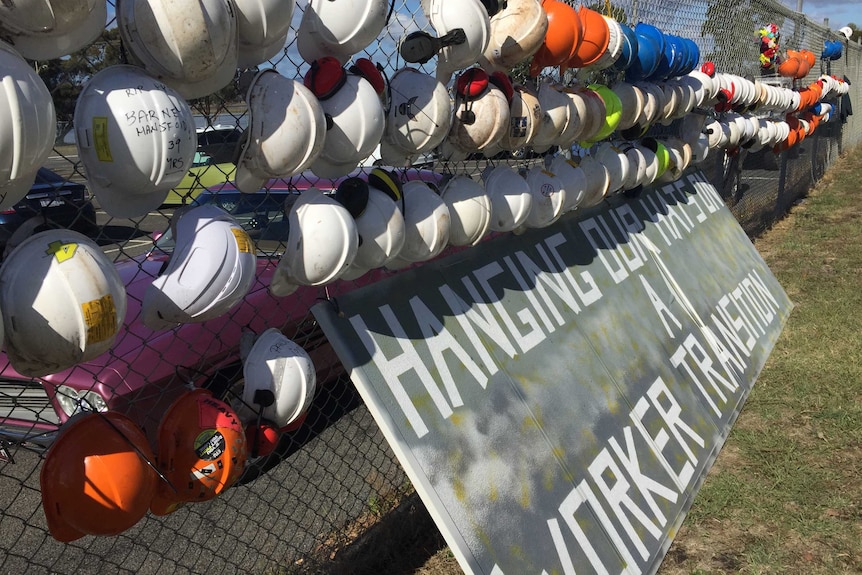 Dozens of hard hats hang on a cyclone fence, above a sign reading "hanging out hats on a worker transition".