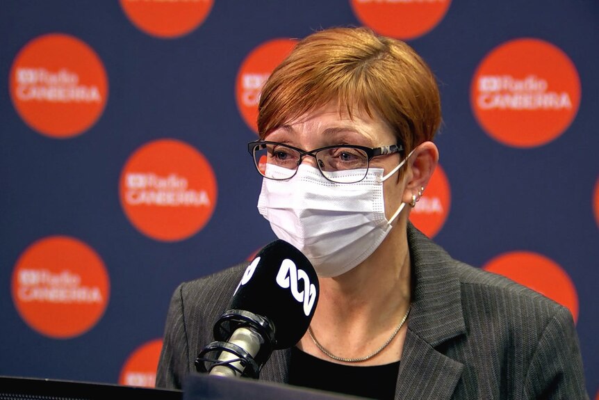 A suited woman with glasses wearing a white mask sits in front of a microphone.