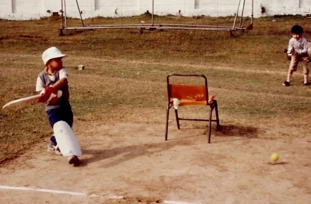 Two little boys play cricket outdoors on a dirt field. One boy bats the other fields. A chair is in the centre.