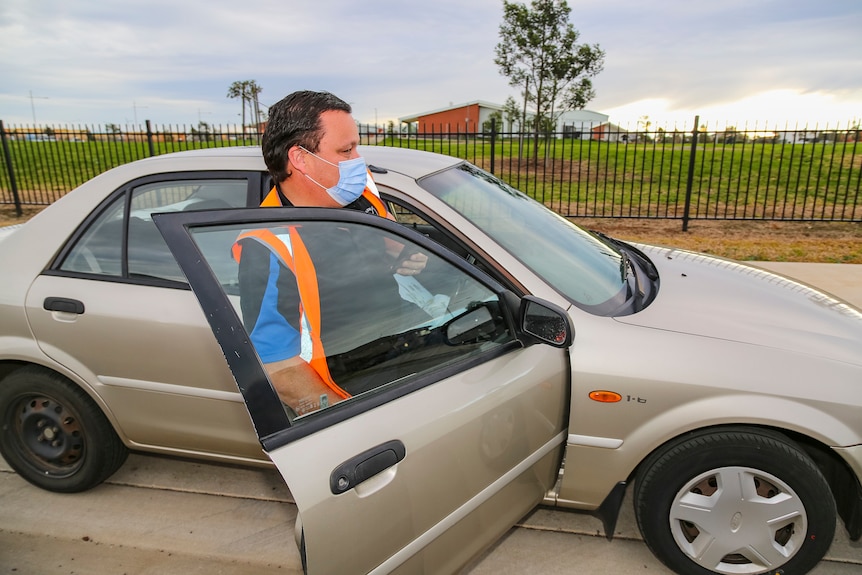 A man in an orange high-vis vest and surgical face mask gets back into his car.
