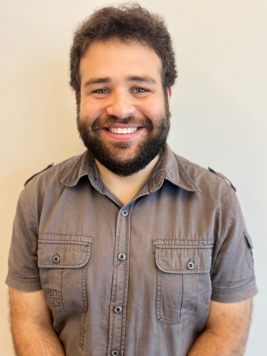 A smiling portrait photo of a young, bearded male teacher in a short-sleeved, grey, collared shirt.