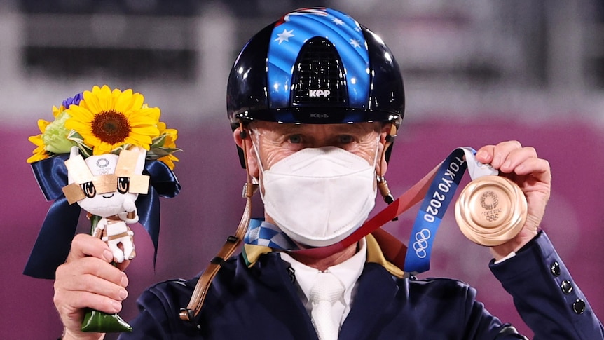 An Australian male competitor in the Tokyo Olympics equestrian competition wears a mask as he holds up his bronze medal.