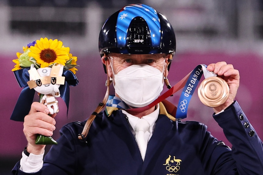 An Australian male competitor in the Tokyo Olympics equestrian competition wears a mask as he holds up his bronze medal.