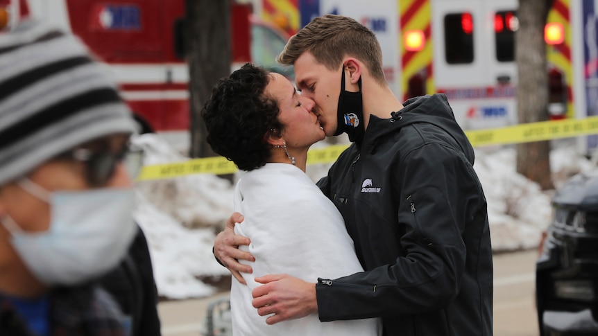 A man and woman kiss with emergency vehicles in the background.