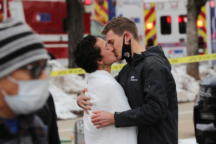 A man and woman kiss with emergency vehicles in the background.
