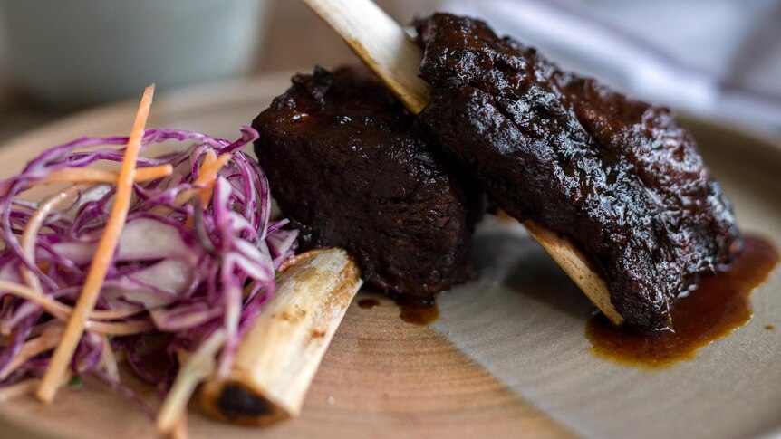Beef short ribs sit on wooden plate with slaw and beer in background 