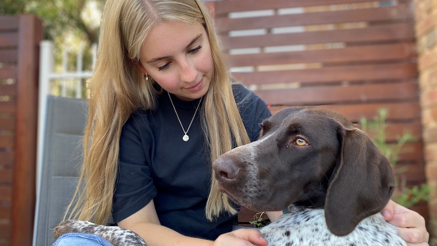 A young woman with blonde hair and black tshirt sits and pats a brown dog 
