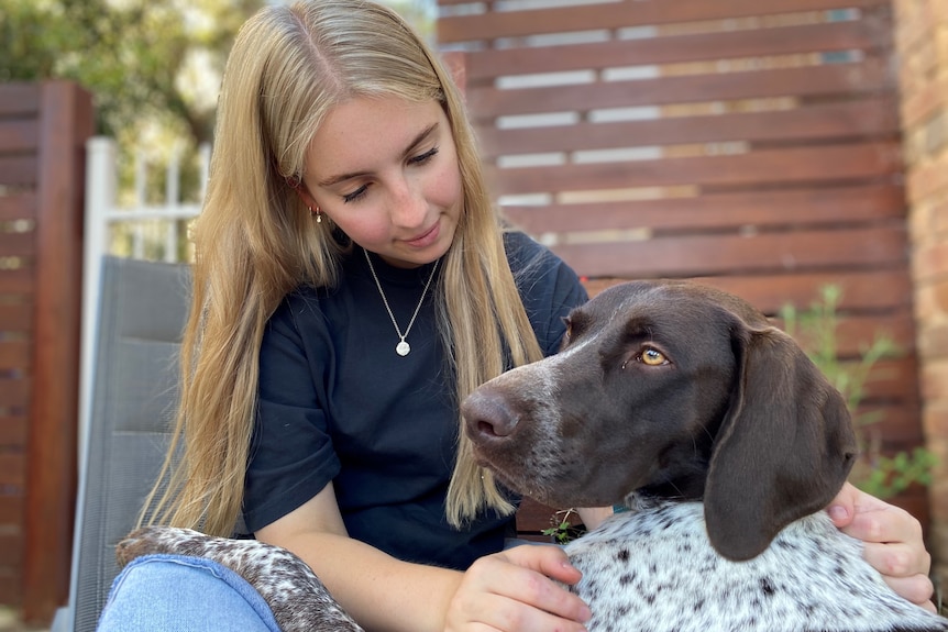 A young woman with blonde hair and black tshirt sits and pats a brown dog 