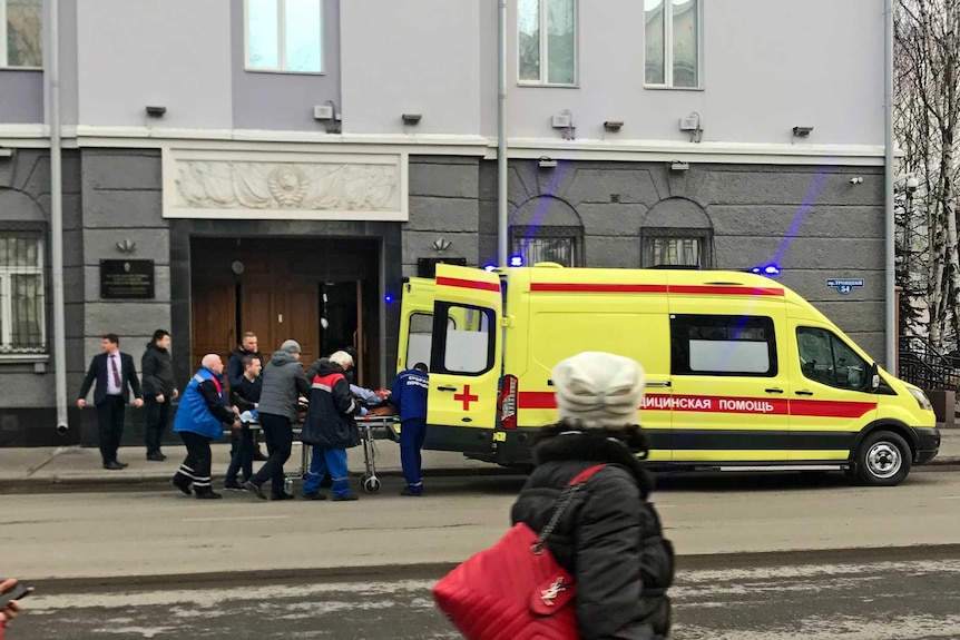 Five people gathered around a stretcher carrying a wounded person load the stretcher into an ambulance, outside a building.