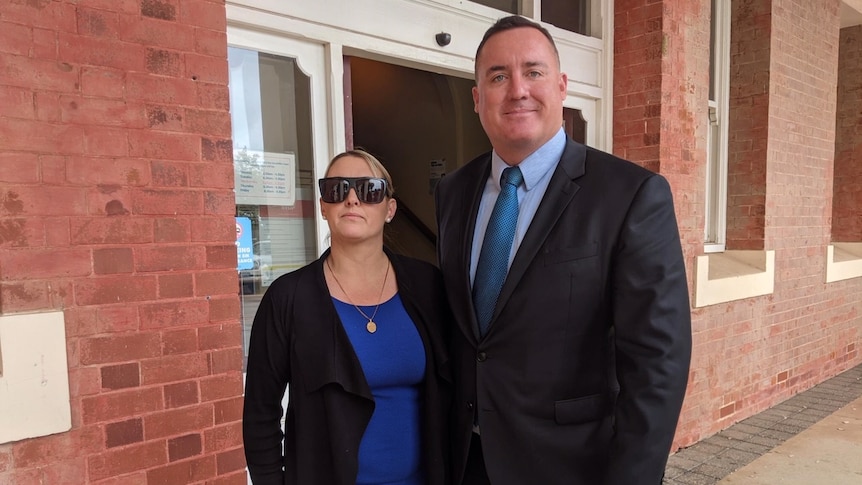 Lady wearing blue dress, black jacket and sunglasses stands beside man in suit.