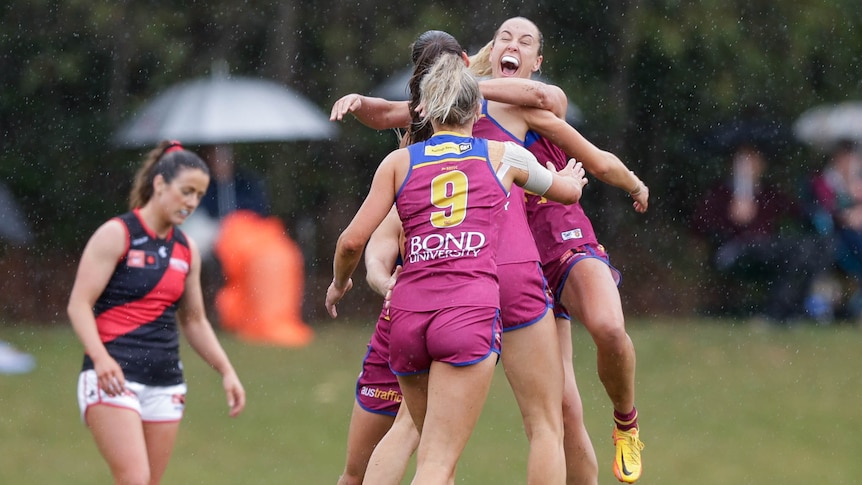 A group of Brisbane Lions AFLW players embrace in the rain, as a dejected Essendon player stands in the background.