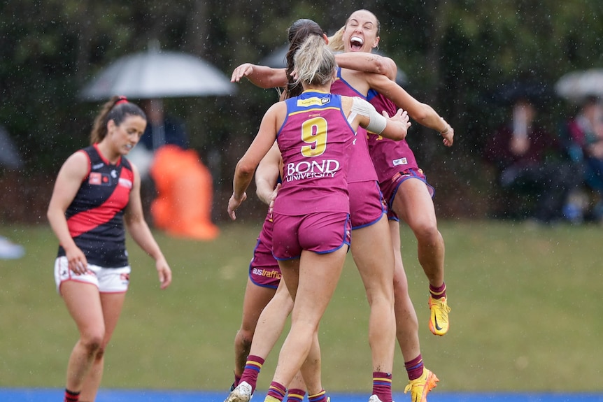 A group of Brisbane Lions AFLW players embrace in the rain, as a dejected Essendon player stands in the background.