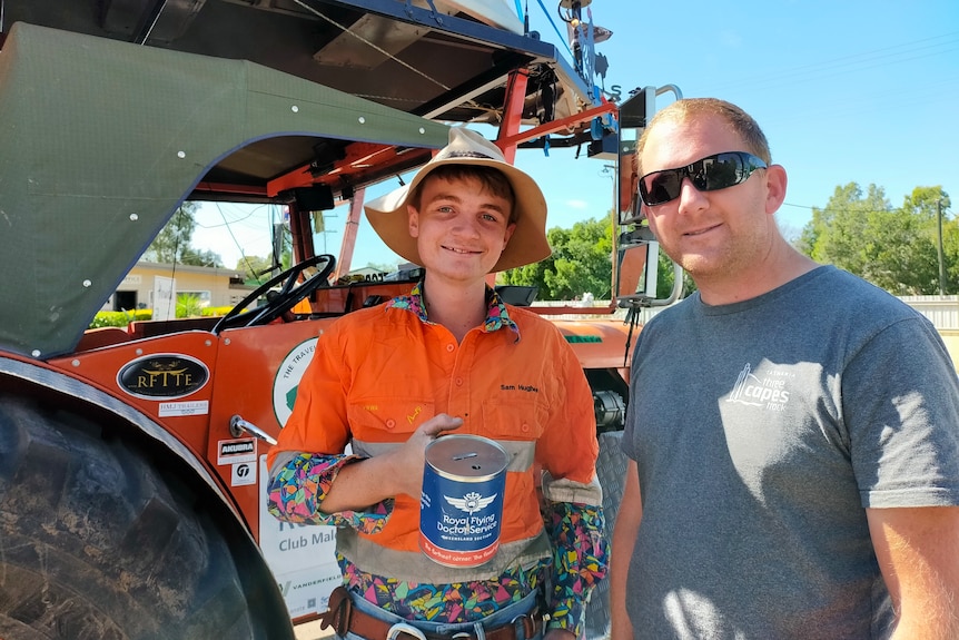 A young man in a bright orange shirt stands in front of a tractor holding a donation tin. A man in a grey shirt is next to him