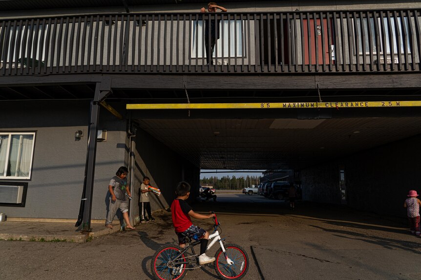 A kid stands on a motel balcony and looks down over a driveway. Other kids on bikes and with water guns are in the driveway.