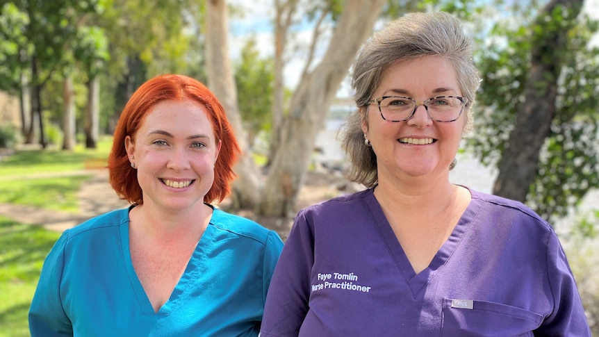Two women in scrubs stand outside, smiling at the camera
