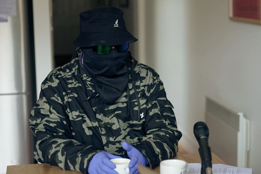 A man covered completely by a hat, sunglasses and mask, sits at a table holding a coffee cup with gloved hands.
