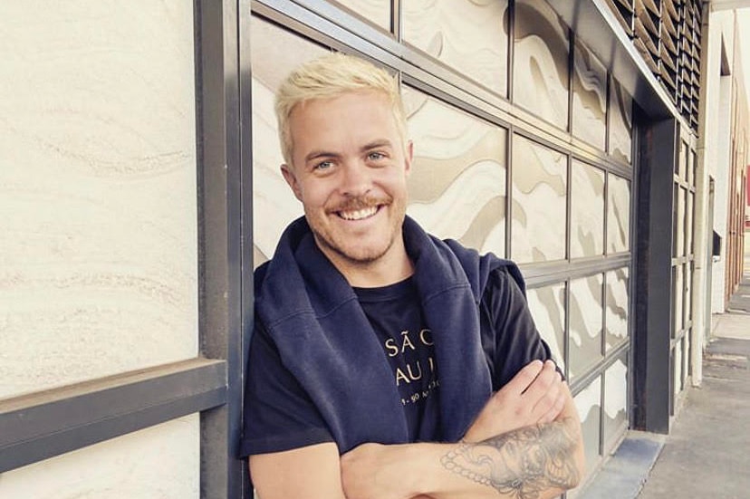 A young man leans against a wall, smiling.