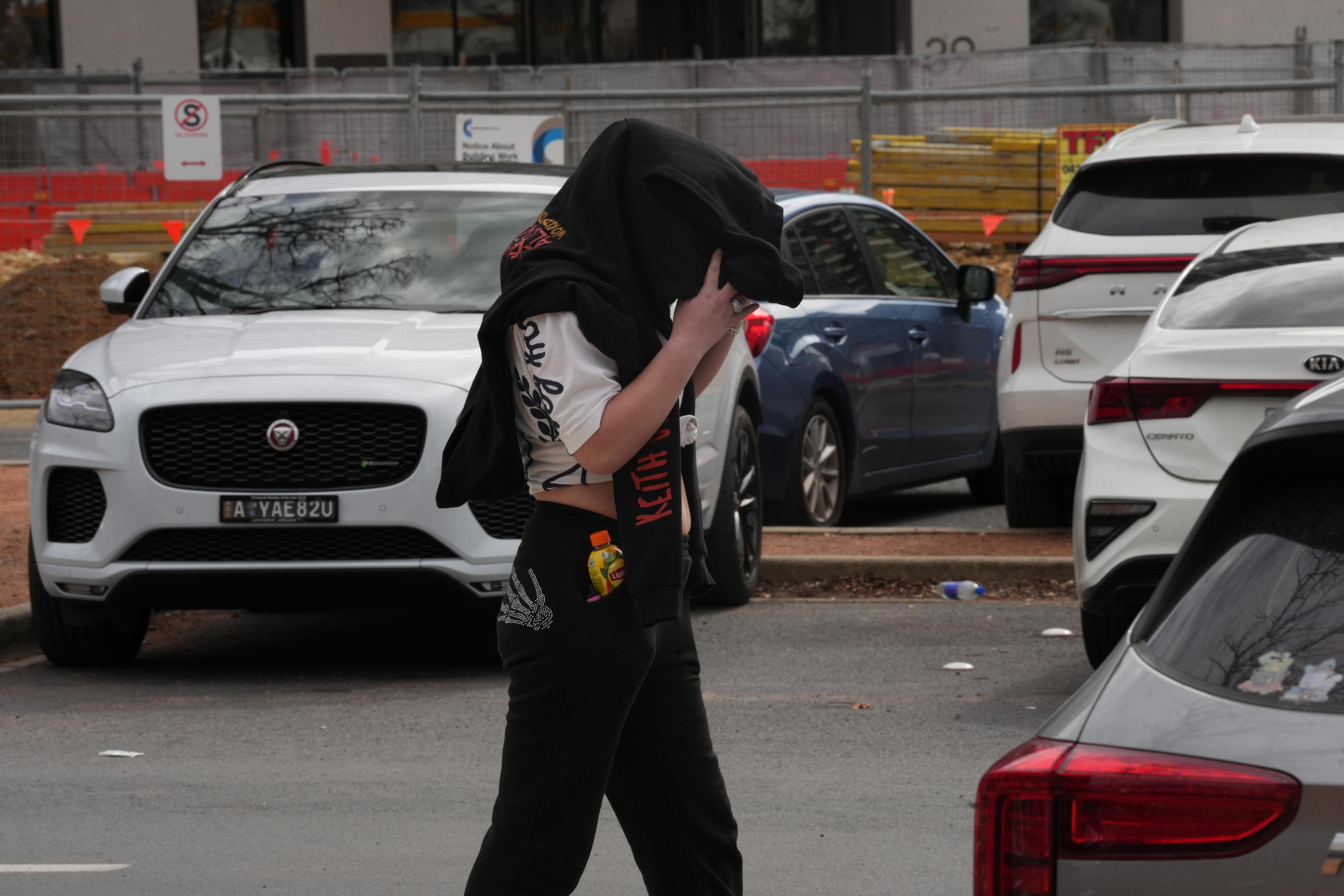 Una persona con una sudadera con capucha negra sobre su cabeza camina por un estacionamiento.