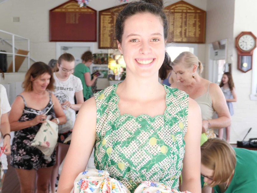 Lily Harrison looks to the camera as she holds to floral bags in her hand. She smiles, and is wearing a green top.