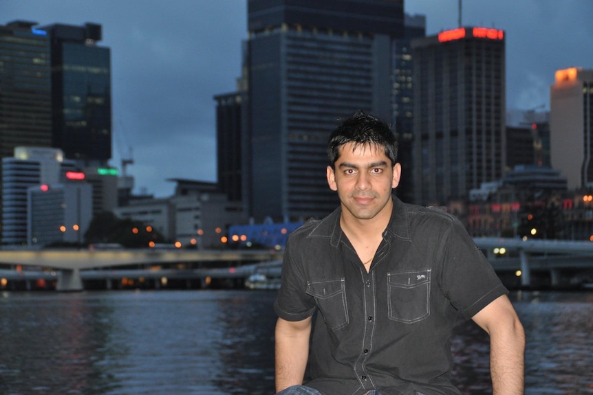 A slightly smiling young man, black hair, black shirt, stands against the Melbourne skyline at dusk.