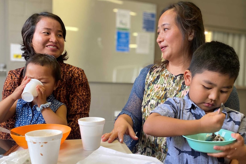 Two women sit at a table with children on their lap. They chat, while children eat and drink.