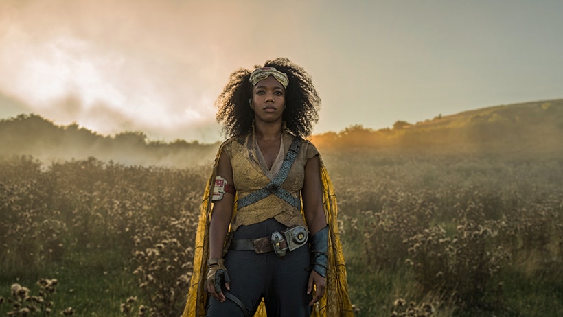 A woman with serious expression wears metal headpiece and cape and stands in field at dusk in front of smokey sky.
