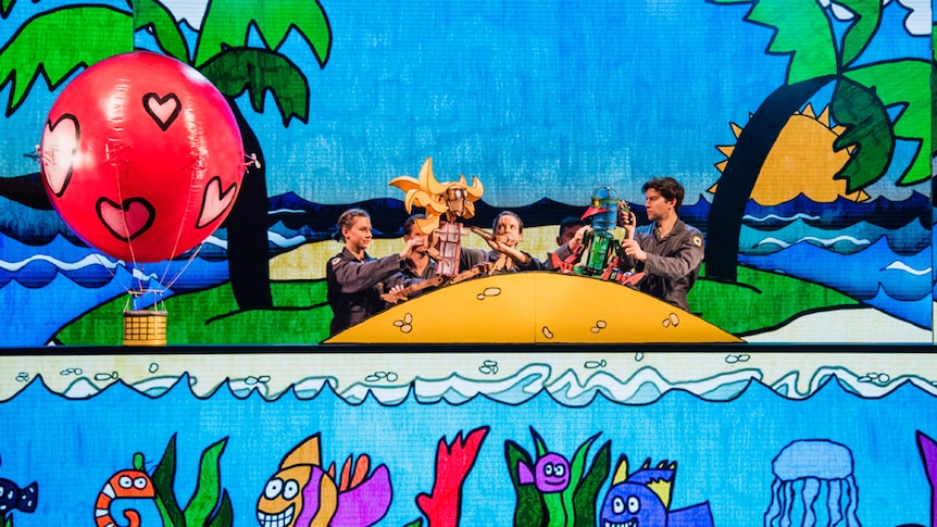 Puppeteers on stage with puppets of Laser Beak Man and another character against a bright animated drawing of a tropical island.