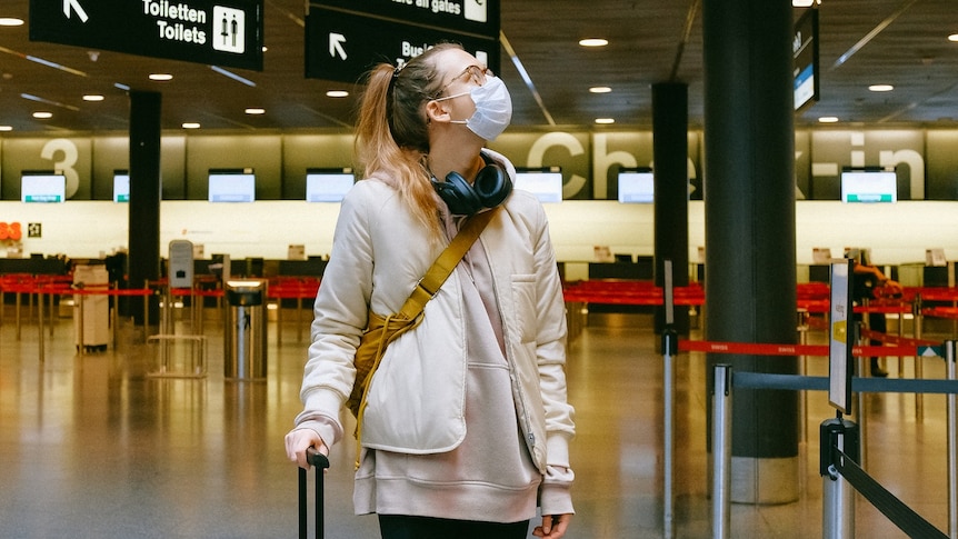 A woman wearing a face mask rests a hand on her suitcase at an airport