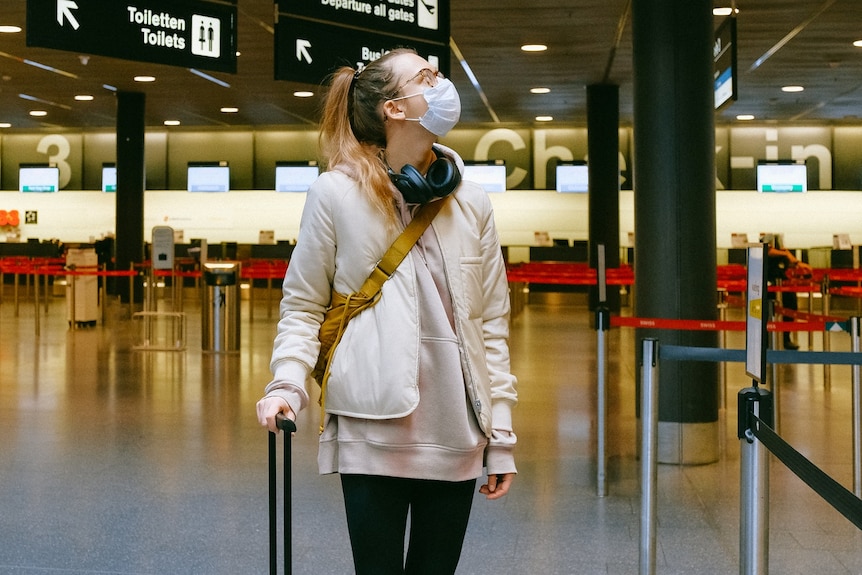 A woman wearing a face mask rests a hand on her suitcase at an airport