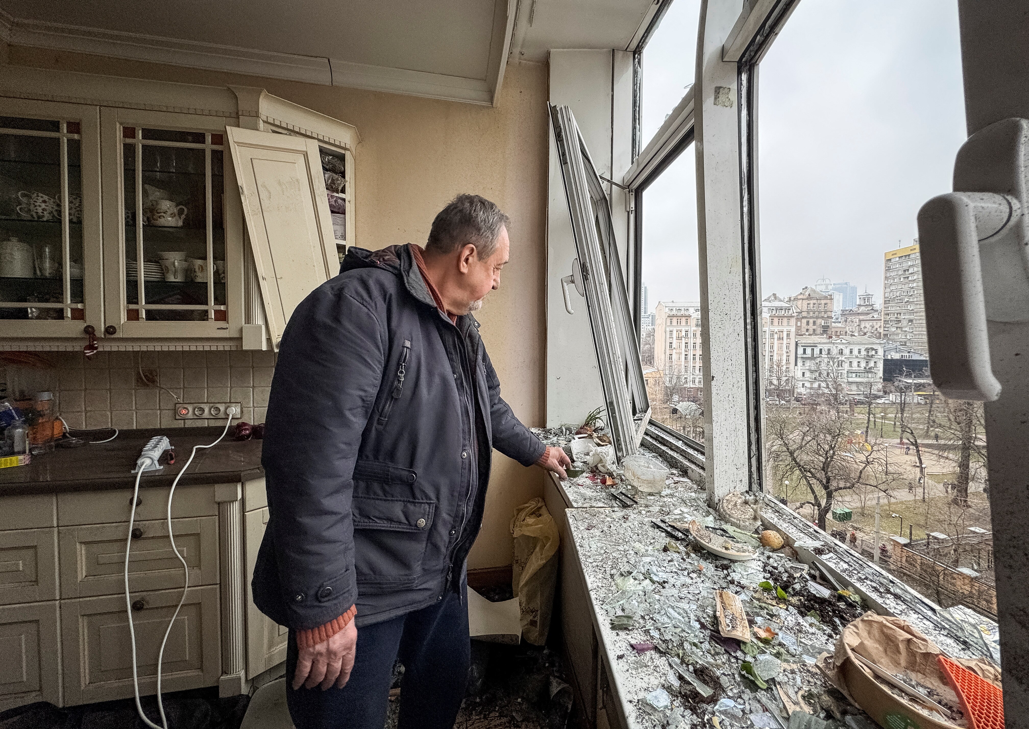 A man stands in his kitchen, which has windows blown out and debris all over the room. 