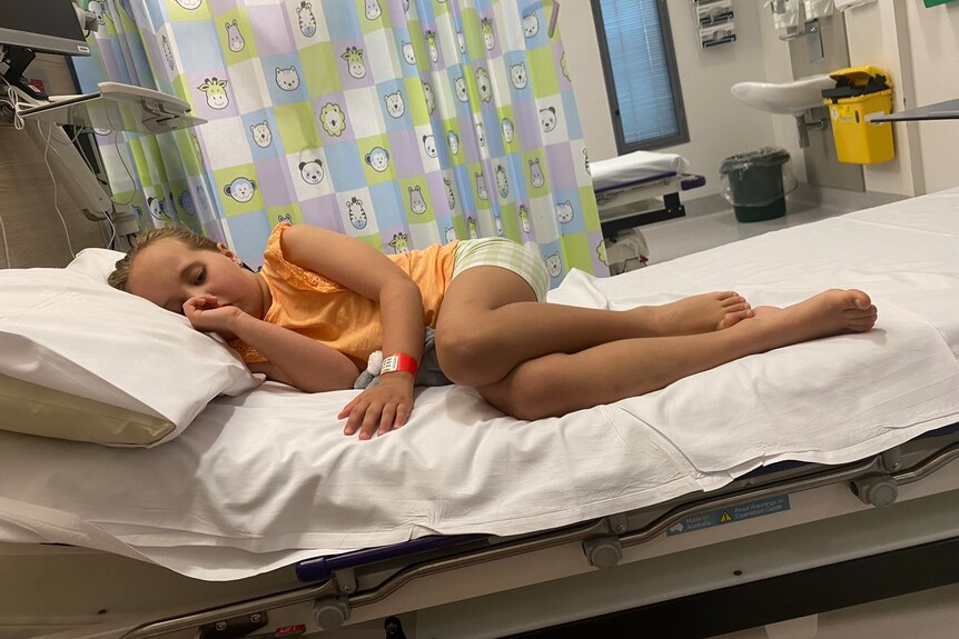 a little girl wearing an orange t shirt and green shorts sleeps in a hospital bed sucking her thumb