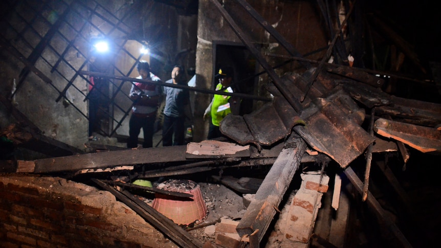 Government officials and a police officer survey a damaged house as wood planks and debris lay scattered across the floor.