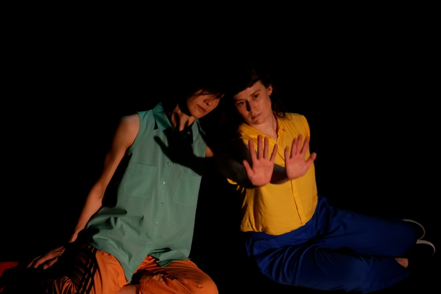 A Japanese woman and an Australian woman sit beside each other, mirroring each other's movements, each with an arm outstretched