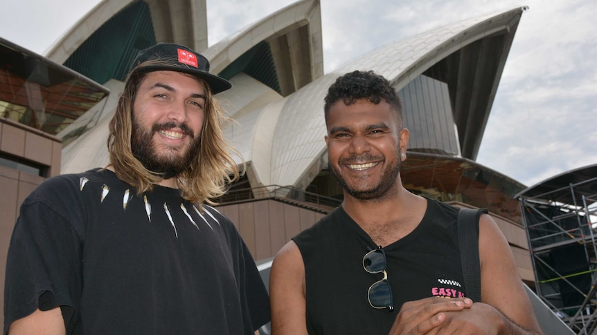 Two men smile as they stand in front of the Sydney Opera House.