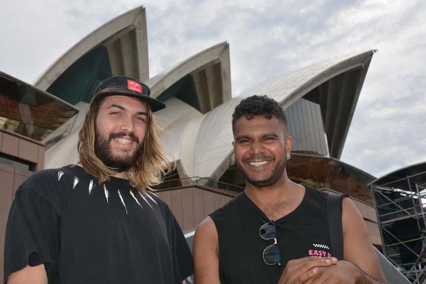 Two men smile as they stand in front of the Sydney Opera House.