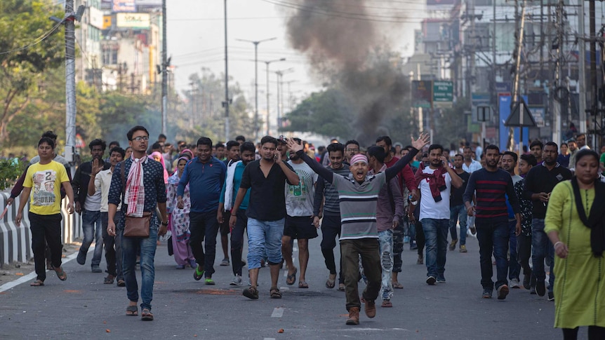 A crowd of protesters walk down a main street. One man screams and holds his hands in the air as smoke rises behind them.