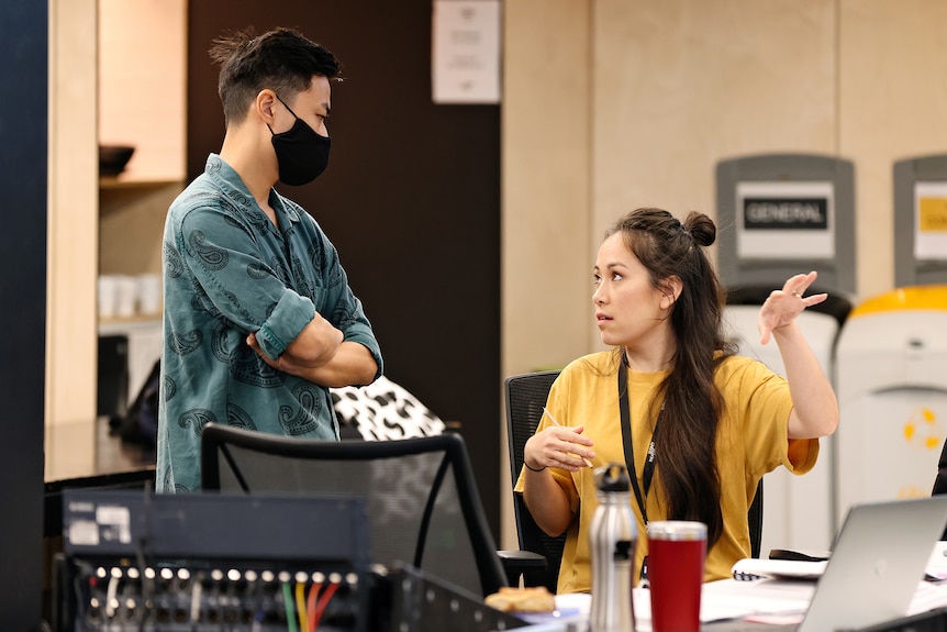 An Asian Australian man stands with his arms folded, wearing a mask, looking at a seated Asian Australian woman who is speaking