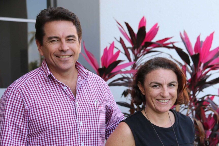 Luke Bowen and Claire George smile at the camera, standing in front of a white wall and a pink plant