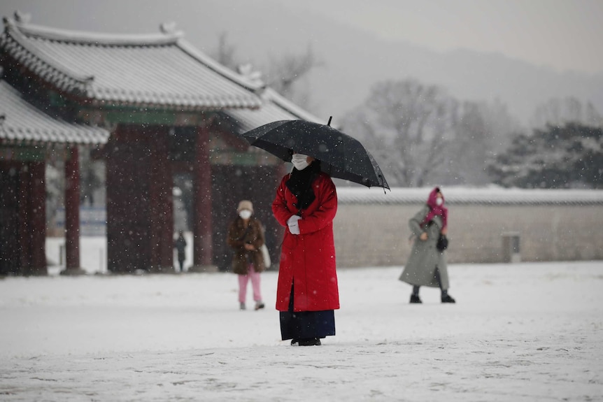 An employee in a red coat wears face mask as a precaution against the coronavirus uses an umbrella to take shelter from the snow