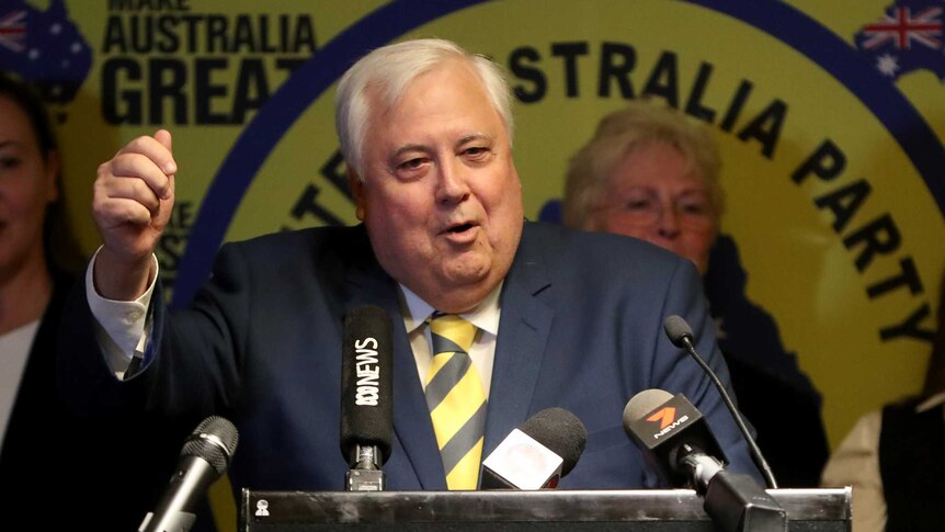 Clive palmer stands at a lectern and makes a gesture with his fist in front of party members and his yellow signage