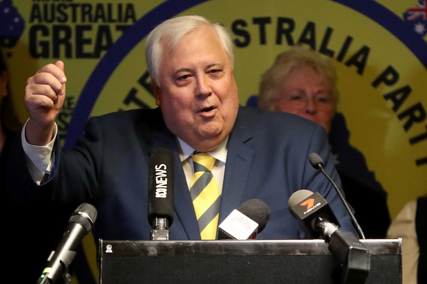 Clive palmer stands at a lectern and makes a gesture with his fist in front of party members and his yellow signage