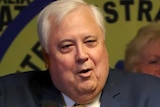 Clive palmer stands at a lectern and makes a gesture with his fist in front of party members and his yellow signage.
