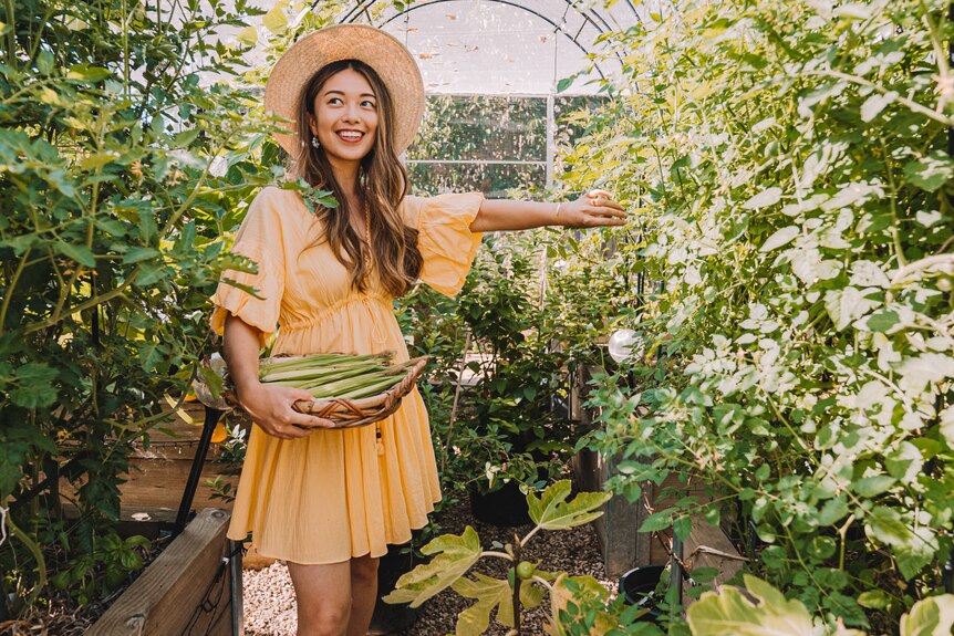 A young woman holds a basket of greens as she stands between rows of raised garden beds under a canopy.