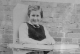 A black and white photo of Lyn Bates standing with assistance from a wooden platform with wheels.