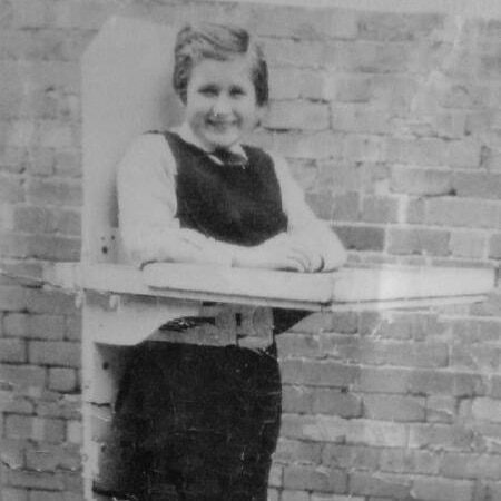 A black and white photo of Lyn Bates standing with assistance from a wooden platform with wheels.