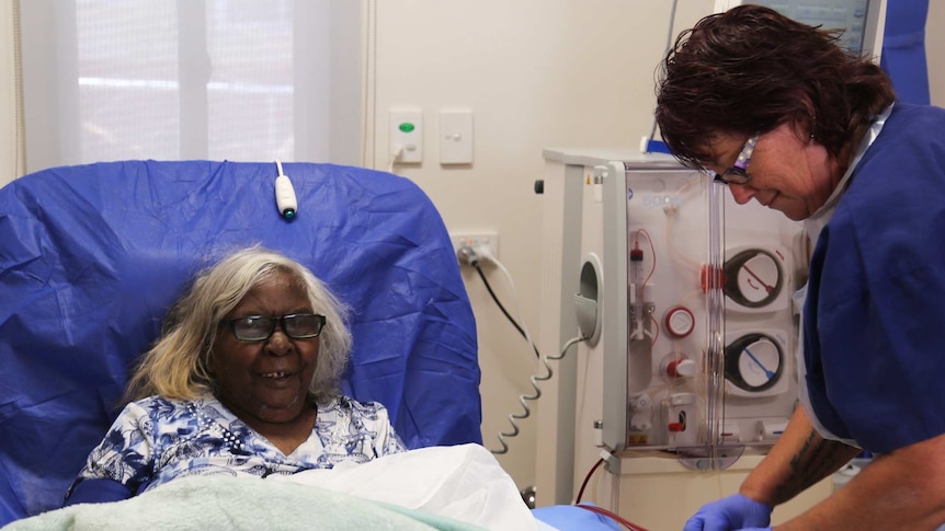 An elderly woman sits upright on a hospital bed as she receives dialysis treatment from a nurse by her bed.