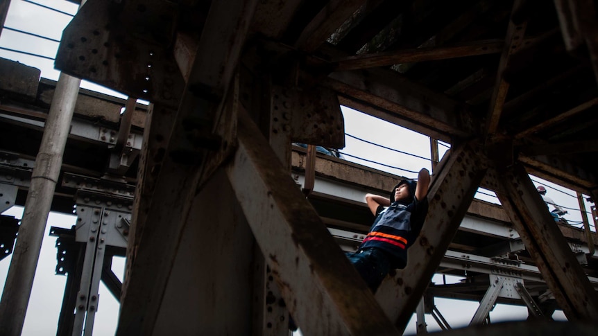 A boy reclines against a rafter under a train bridge